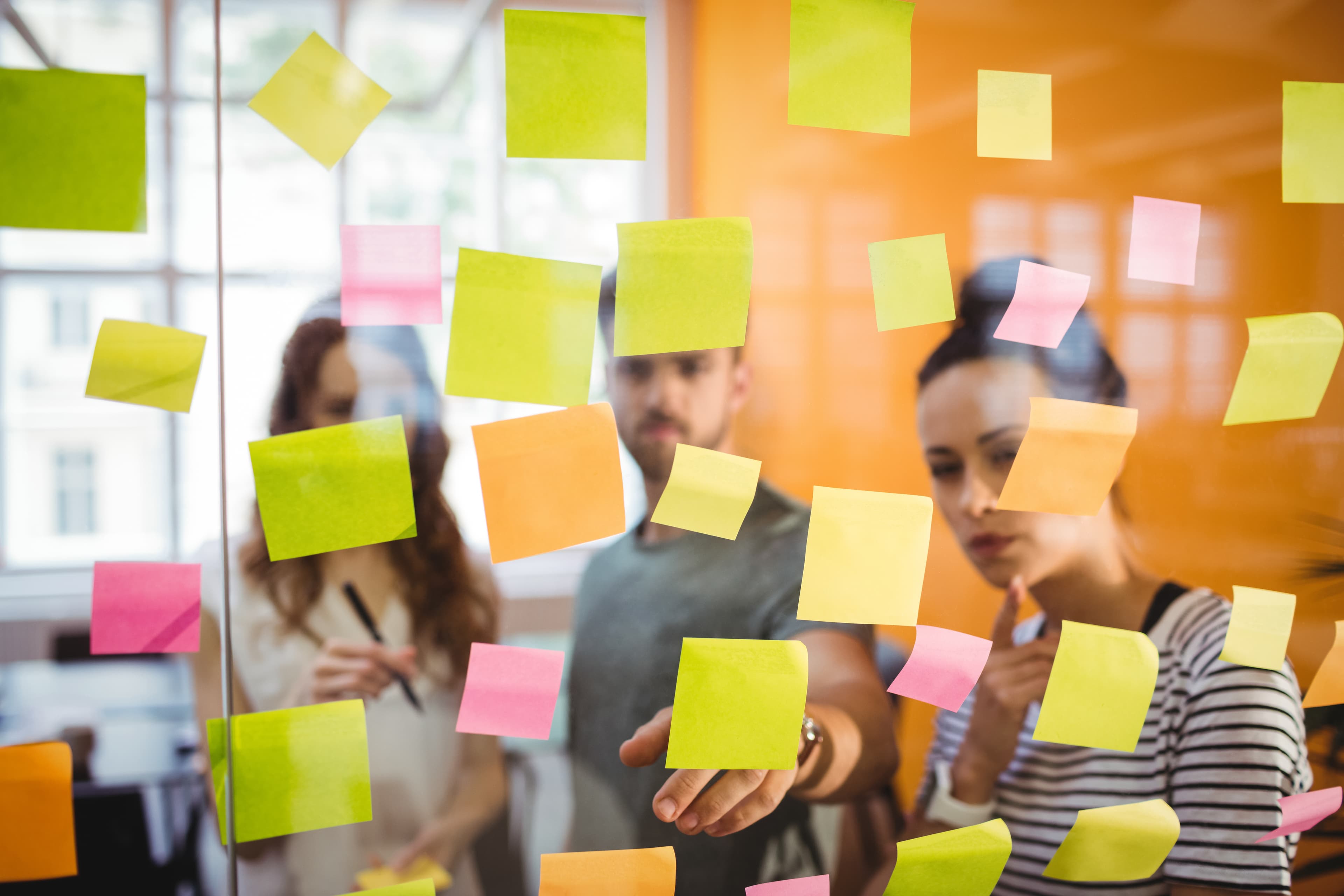 Três pessoas trabalhando em uma sessão de brainstorming. Elas estão analisando uma parede de vidro cheia de post-its coloridos (amarelo, verde, laranja e rosa), enquanto discutem e organizam ideias. A expressão concentrada das pessoas e a variedade de notas indicam um ambiente de colaboração e criatividade.